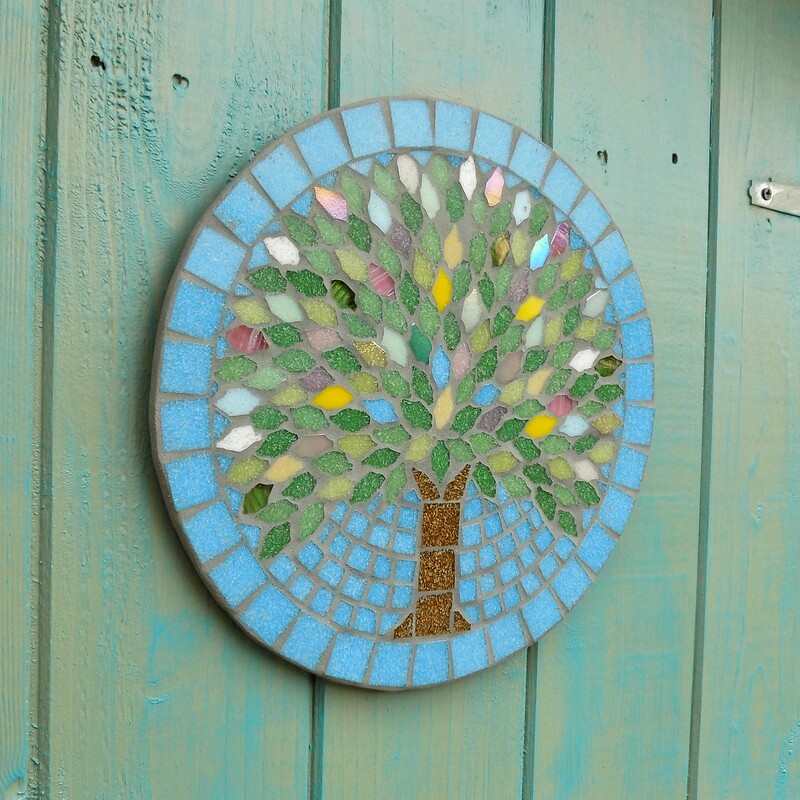 A mosaic garden plaque with a design of a spring tree with leaves made in the colours of spring blossom and flowers on a turquoise blue background