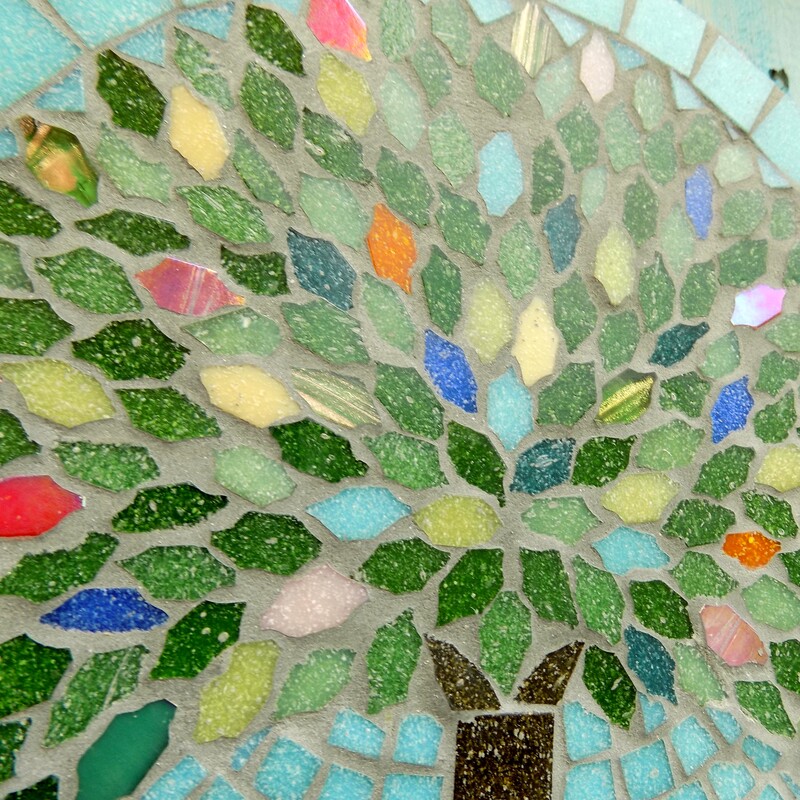 A handmade mosaic garden plaque with a design of a summer tree with leaves made in the colours of rich summer flowers and deep green foliage on a pastel sea green background