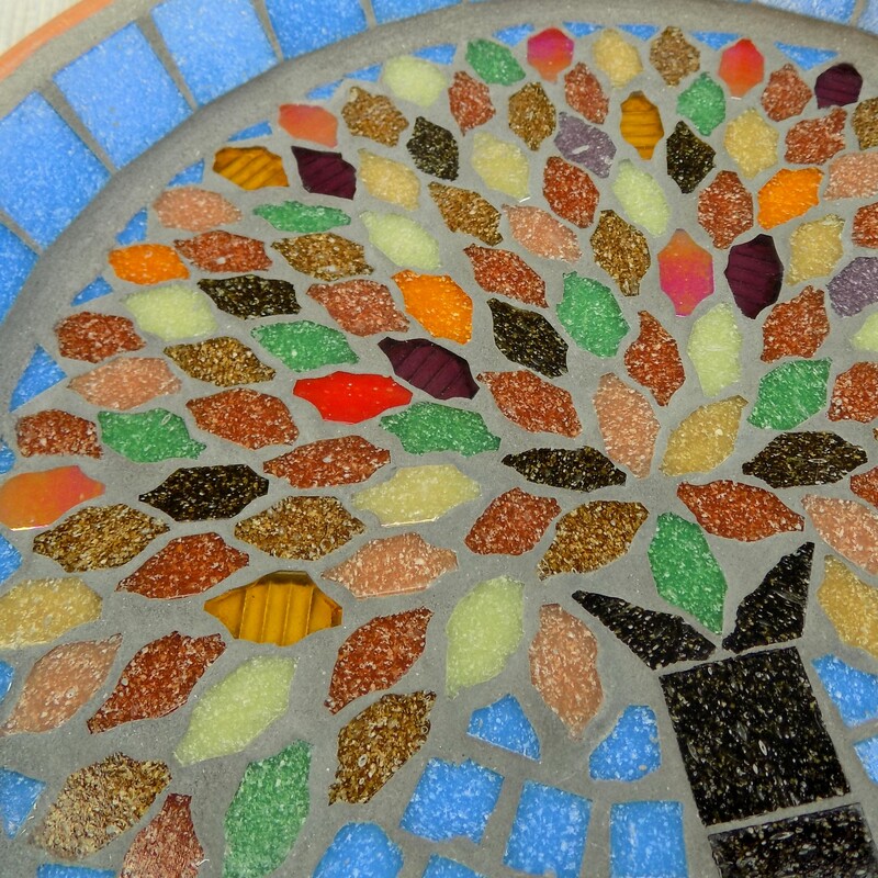 A mosaic birdbath with a design of an tree with autumn colours used in the tile leaves on a sky blue background