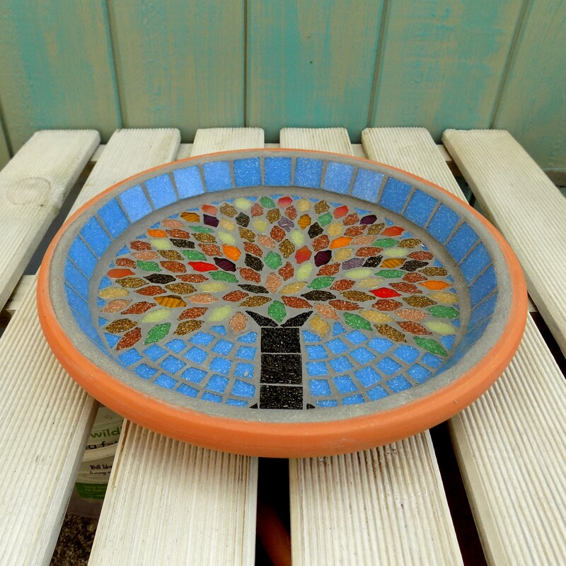A mosaic bird bath with a design of an tree with autumn colours used in the tile leaves on a sky blue background