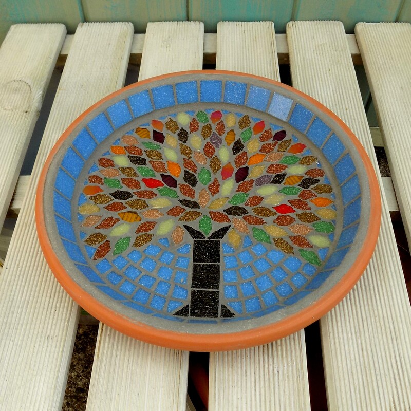 A mosaic garden bird bath with a design of an tree with autumn colours used in the tile leaves on a sky blue background