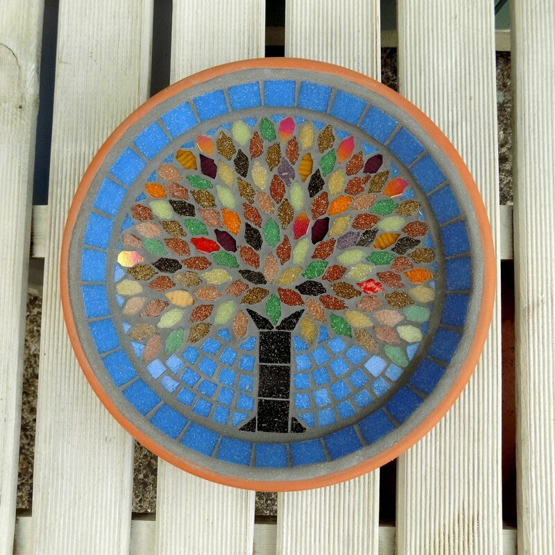 A mosaic garden birdbath with a design of an tree with autumn colours used in the tile leaves on a sky blue background