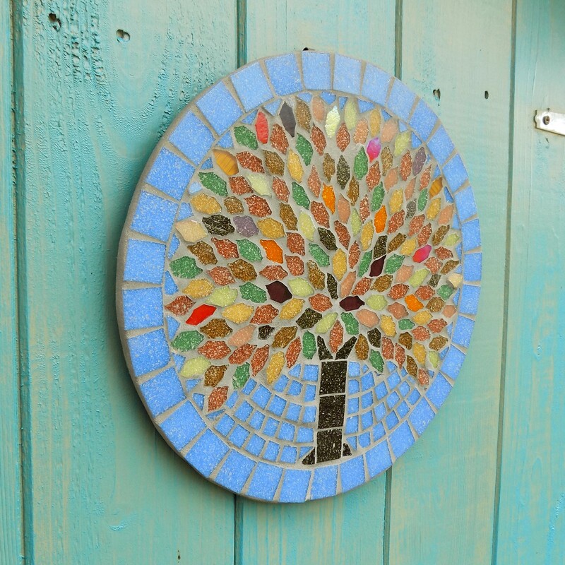 A mosaic garden hanging plaque with a tree design using the rich colours of autumn in the leaves on a sky blue background