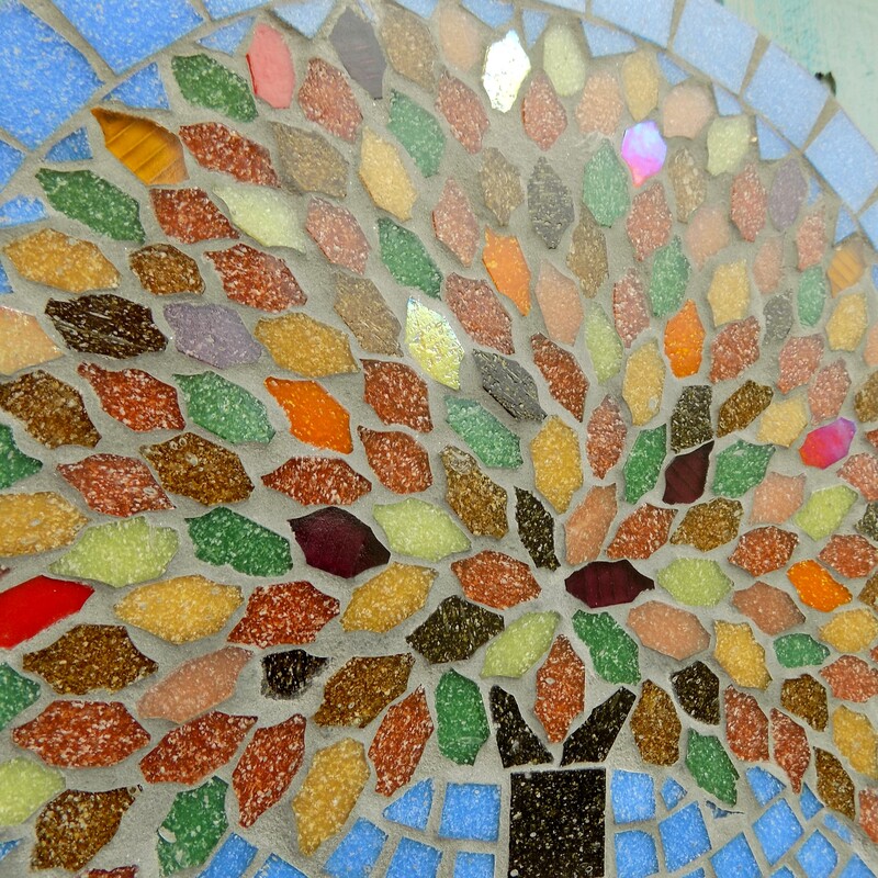 A handmade mosaic garden hanging plaque with a tree design using the rich colours of autumn in the leaves on a sky blue background