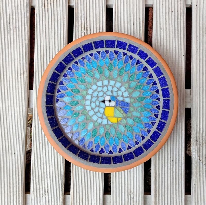 A mosaic bird bath with a bluetit bird in the centre of a splash effect made with blues, turquoise and greens.