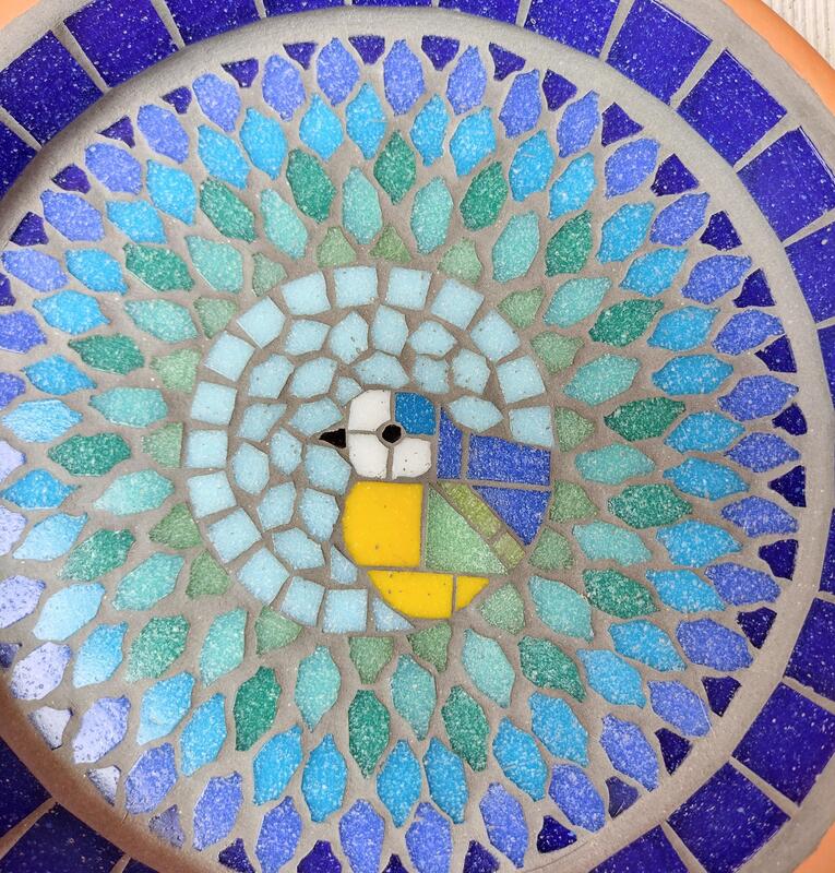 A mosaic birdbath with a bluetit bird in the centre of a splash effect made with blues, turquoise and greens.