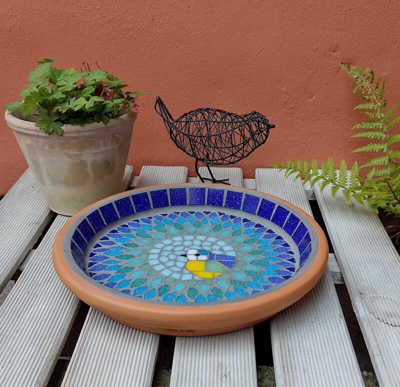 A mosaic garden bird bath with a bluetit bird in the centre of a splash effect made with blues, turquoise and greens.