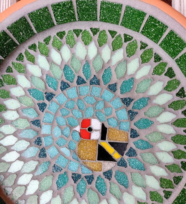 A mosaic garden bird bath with a design of a goldfinch bird in the centre of a splash effect of green tiles