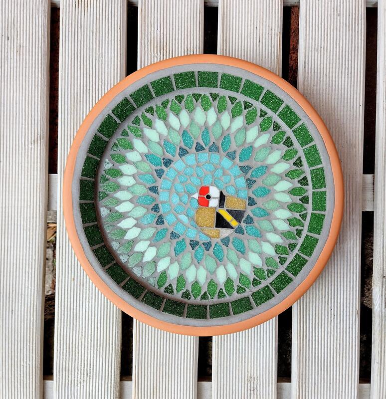 A mosaic bird bath with a design of a goldfinch bird in the centre of a splash effect of green tiles