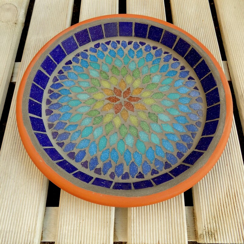 A mosaic bird bath with a mandala style design in the colours of a rainbow from terracotta red in the centre to indigo blue on the rim