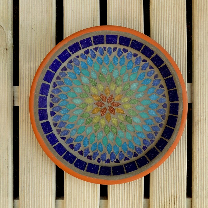 A mosaic garden bird bath with a mandala style design in the colours of a rainbow from terracotta red in the centre to indigo blue on the rim