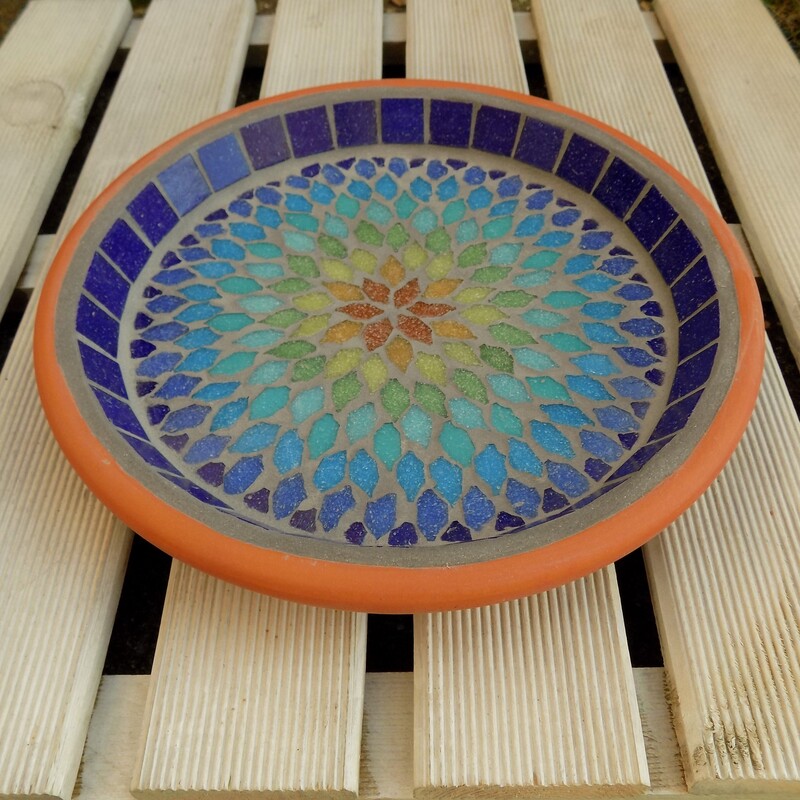 A mosaic garden birdbath with a mandala style design in the colours of a rainbow from terracotta red in the centre to indigo blue on the rim