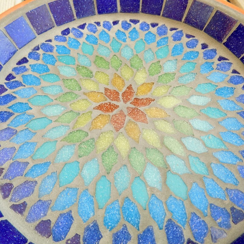 A mosaic birdbath with a mandala style design in the colours of a rainbow from terracotta red in the centre to indigo blue on the rim