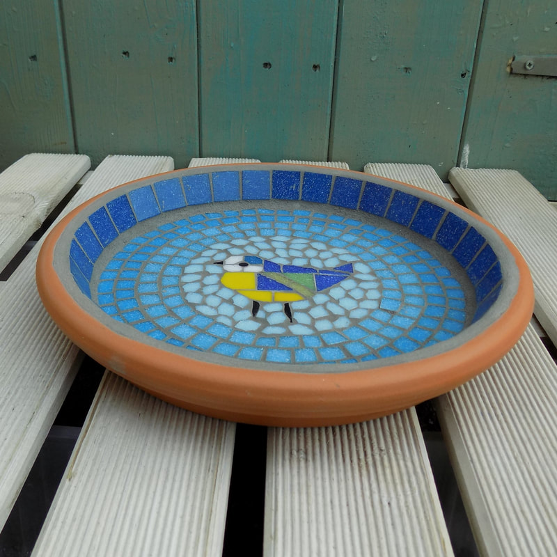 A mosaic birdbath with a design of a bluetit bird in the centre of graduating shades of rings of turquoise tiles from dark on the border to a light circle of aqua around the bird
