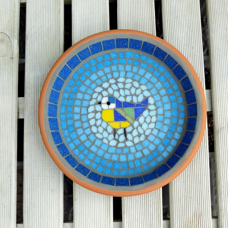 A mosaic garden birdbath with a design of a bluetit bird in the centre of graduating shades of rings of turquoise tiles from dark on the border to a light circle of aqua around the bird