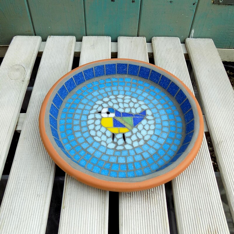 A mosaic garden bird bath with a design of a bluetit bird in the centre of graduating shades of rings of turquoise tiles from dark on the border to a light circle of aqua around the bird