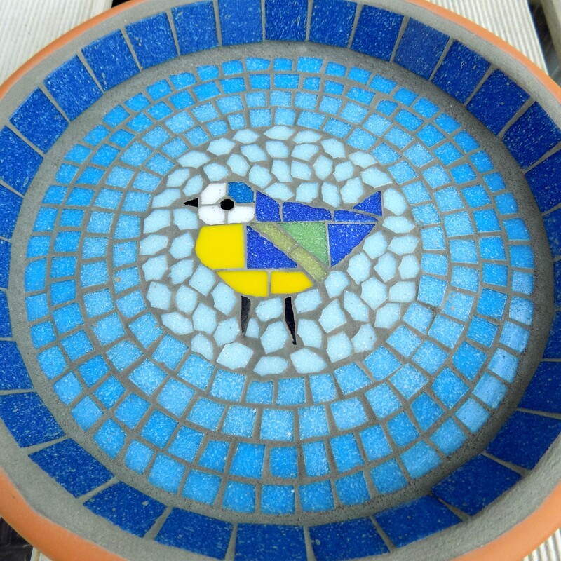 A mosaic birdbath with a design of a blue tit bird in the centre of graduating shades of rings of turquoise tiles from dark on the border to a light circle of aqua around the bird