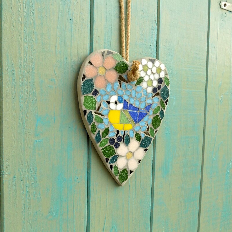 A mosaic hanging heart with a design of a bluetit bird standing surrounded by a border of hedgerow leaves and flowers