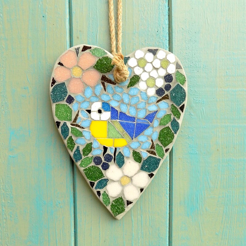 A mosaic hanging heart with a design of a blue tit bird standing surrounded by a border of hedgerow leaves and flowers