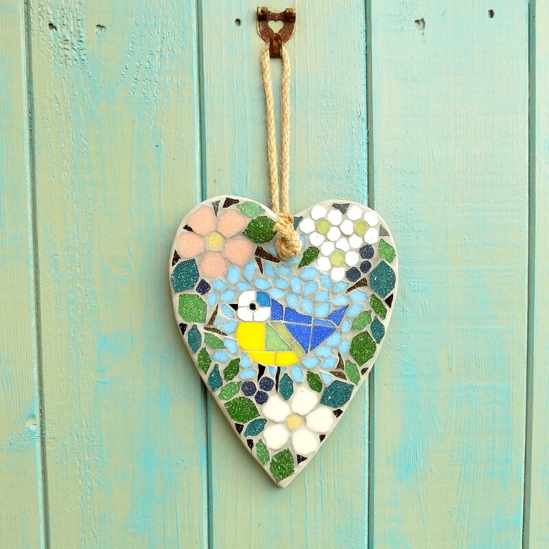 A mosaic garden hanging heart with a design of a bluetit bird standing surrounded by a border of hedgerow leaves and flowers