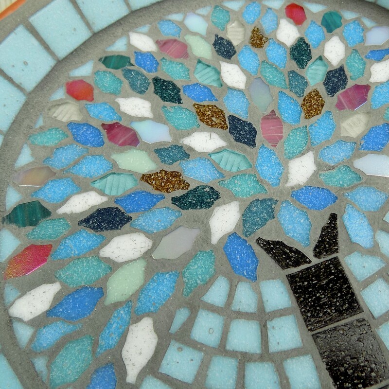A mosaic garden bird bath with a design of a tree with leaves made using the colours of winter snow, ice and sunsets on a pale aqua background