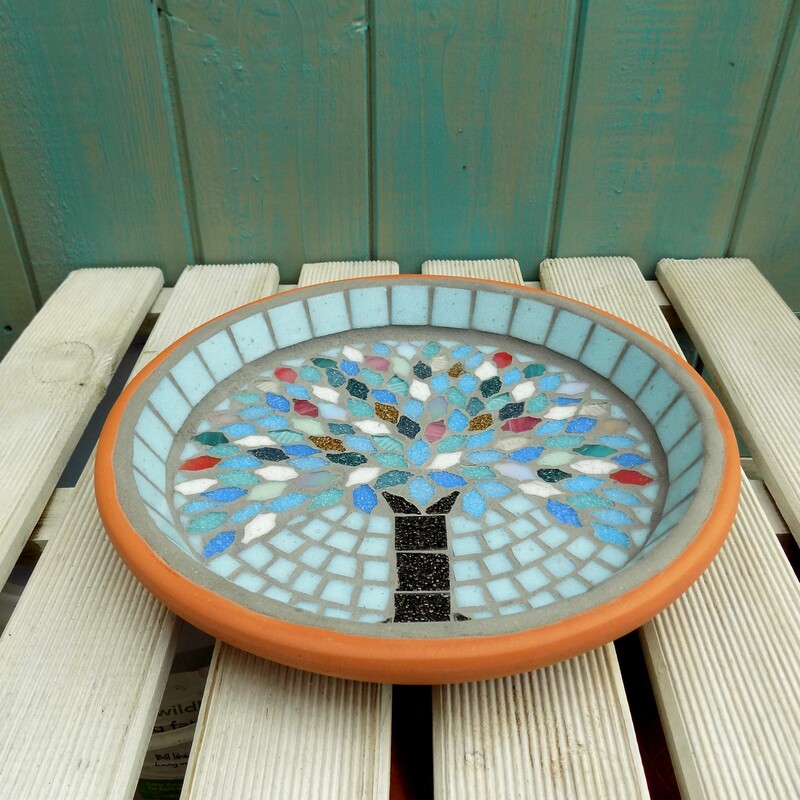 A mosaic bird bath with a design of a tree with leaves made using the colours of winter snow, ice and sunsets on a pale aqua background