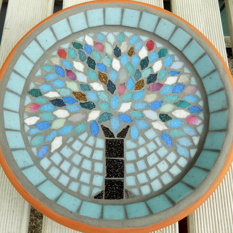 A mosaic birdbath with a design of a tree with leaves made using the colours of winter snow, ice and sunsets on a pale aqua background