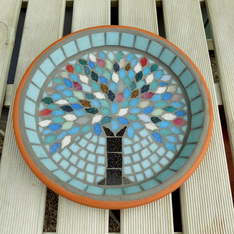 A mosaic garden birdbath with a design of a tree with leaves made using the colours of winter snow, ice and sunsets on a pale aqua background