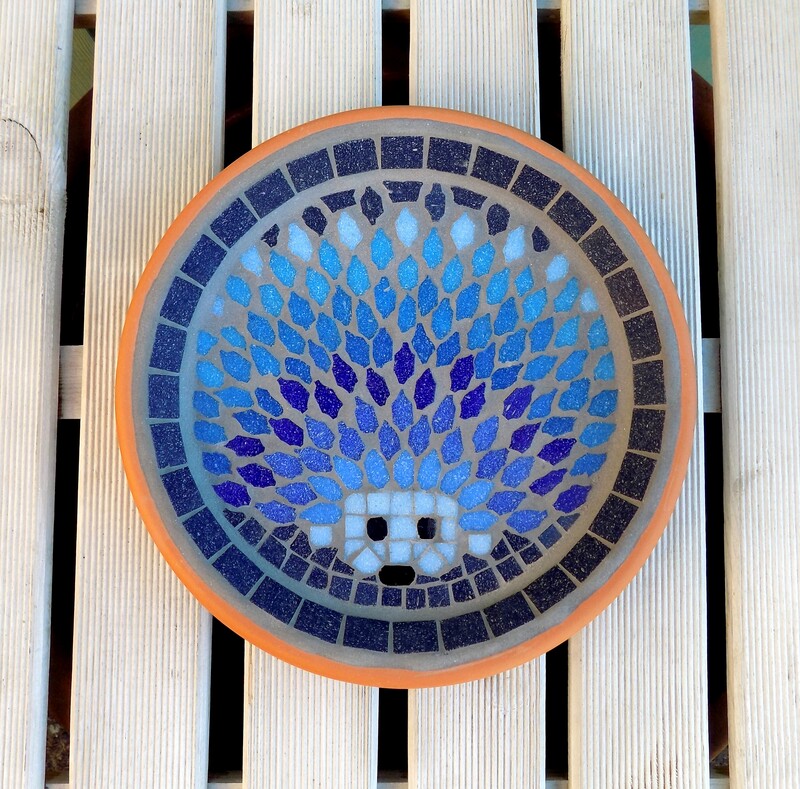 A mosaic garden bird bath with a design of  a hedgehog made in shades of blues with a deep navy background