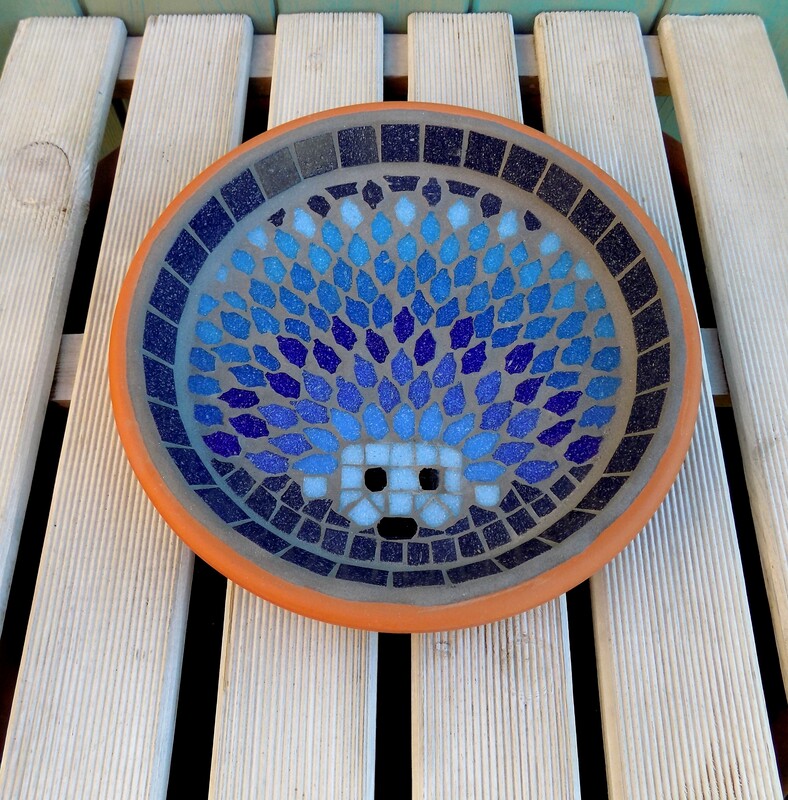 A mosaic garden birdbath with a design of  a hedgehog made in shades of blues with a deep navy background