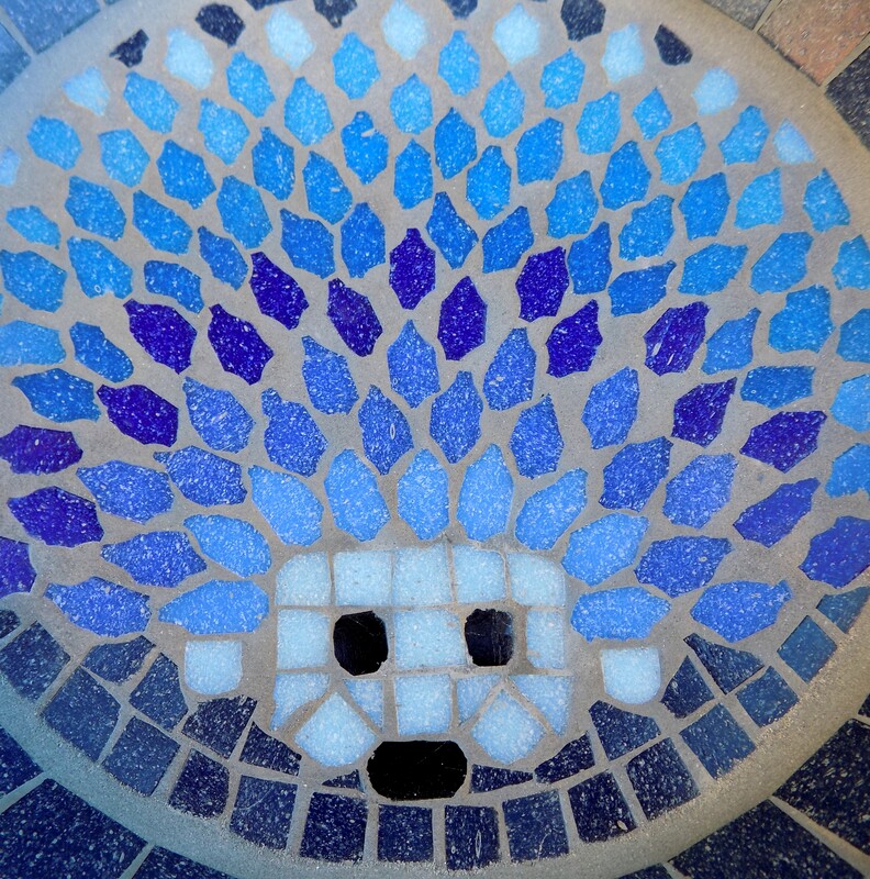 A mosaic bird bath with a design of  a hedgehog made in shades of blues with a deep navy background