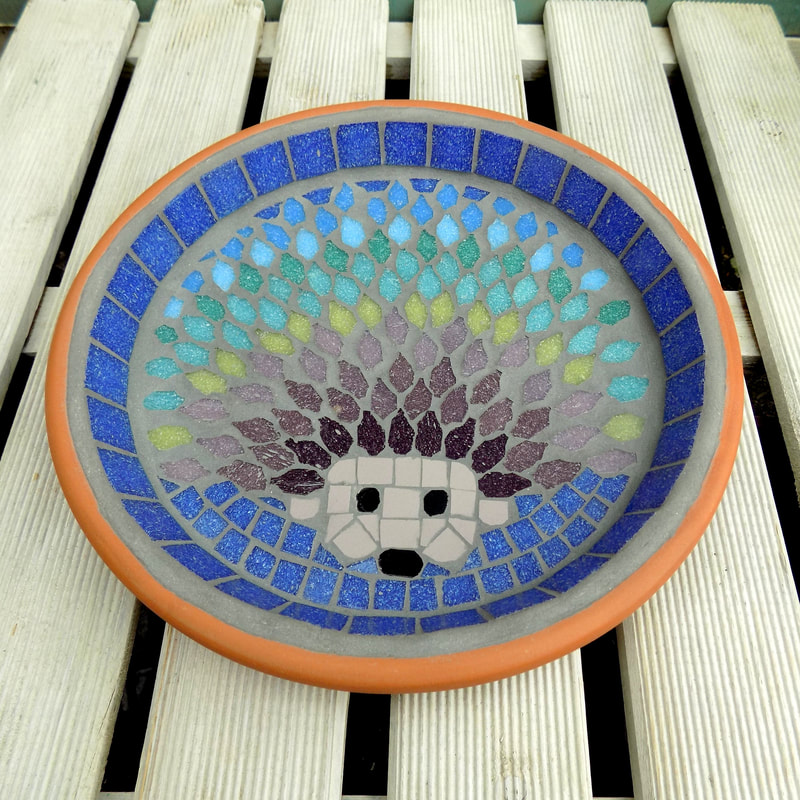 A mosaic garden bird bath with a design of a hedgehog with each line of spines made in a rainbow of tile colours 