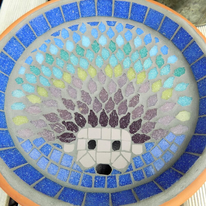 A mosaic bird bath with a design of a hedgehog with each line of spines made in a rainbow of tile colours 
