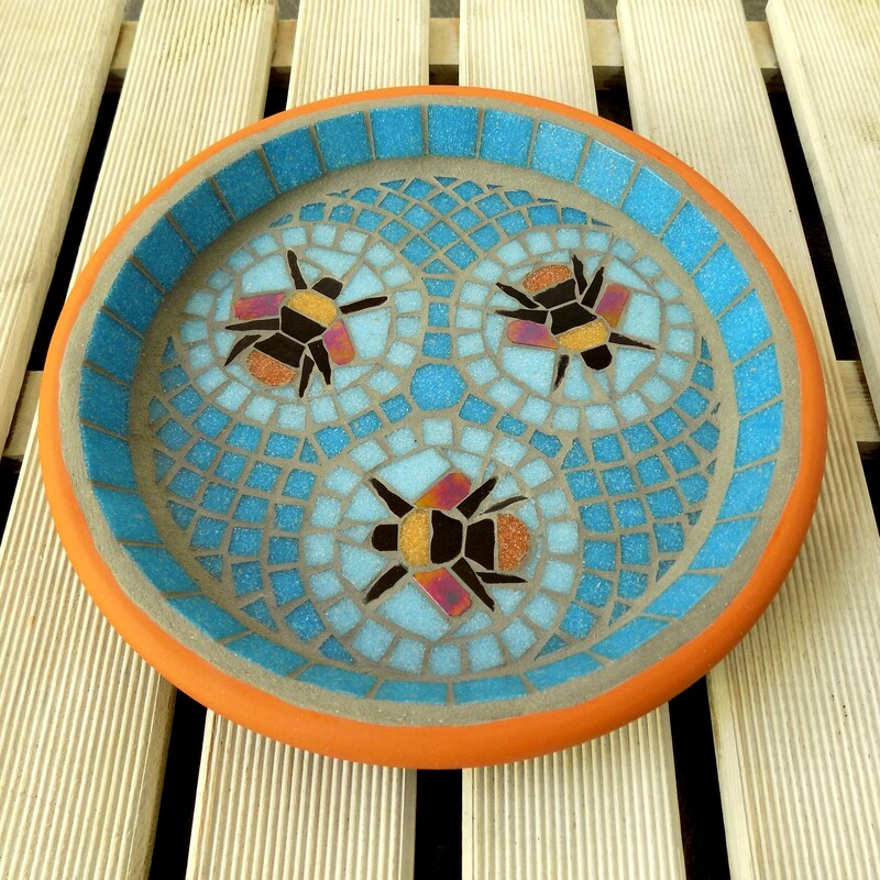 A mosaic bird bath with a design of three bumblebees in aqua blue circles with a geometric pattern of turquoise tiles as a background