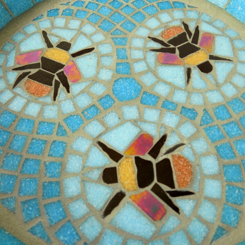 A mosaic birdbath with a design of three bumblebees in aqua blue circles with a geometric pattern of turquoise tiles as a background