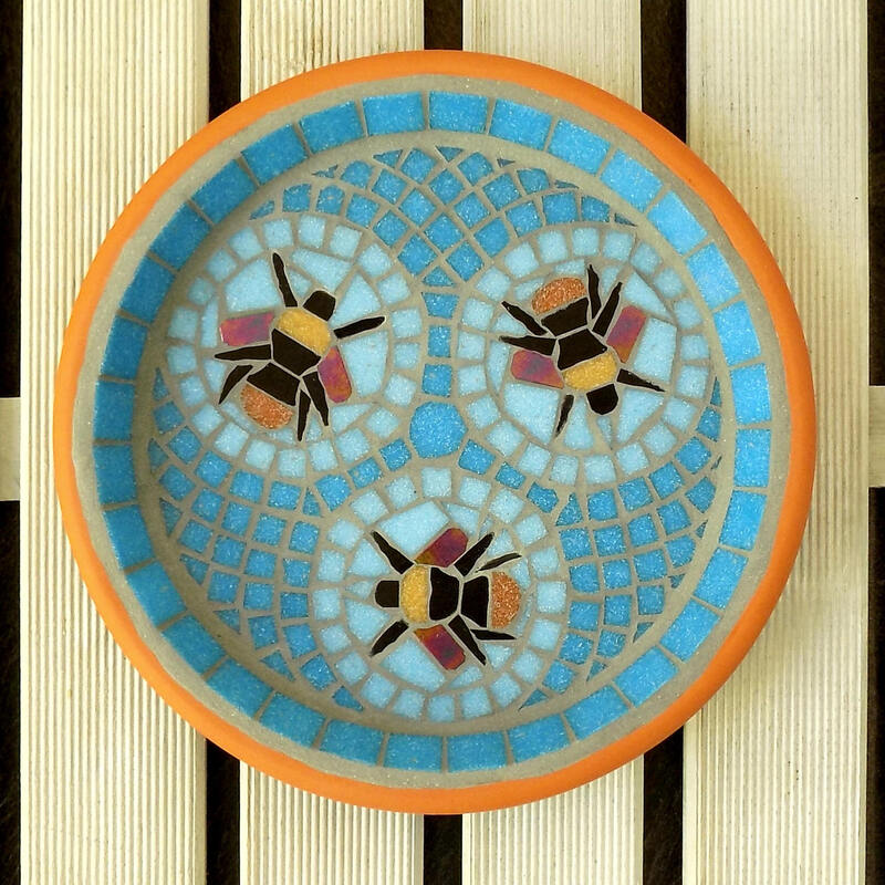 A mosaic garden bird bath with a design of three bumblebees in aqua blue circles with a geometric pattern of turquoise tiles as a background