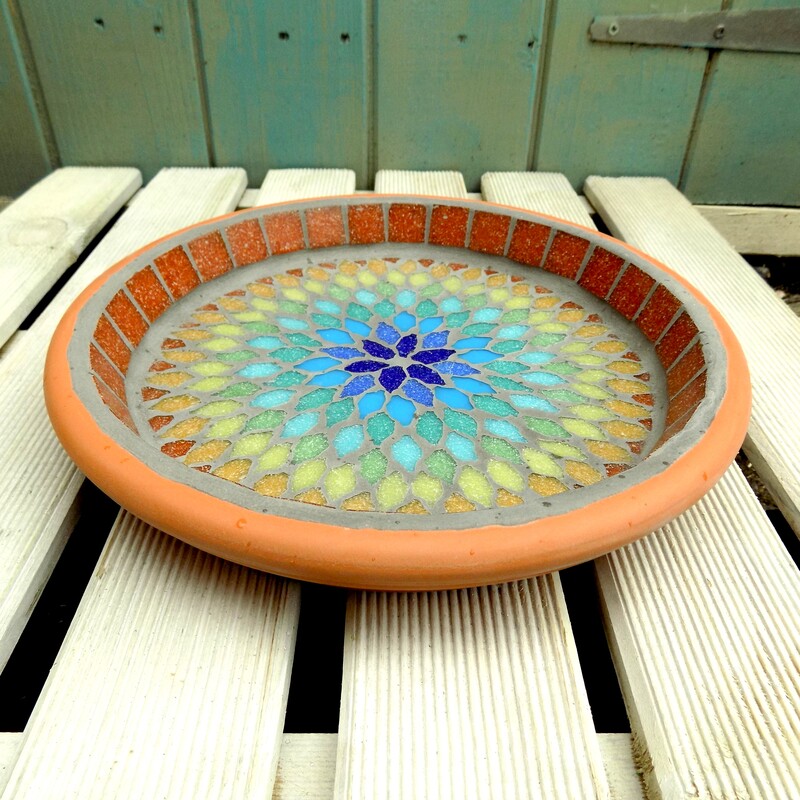 A mosaic birdbath with a  mandala style design in the colours of the rainbow from terracotta on the outside to indigo blue in the centre