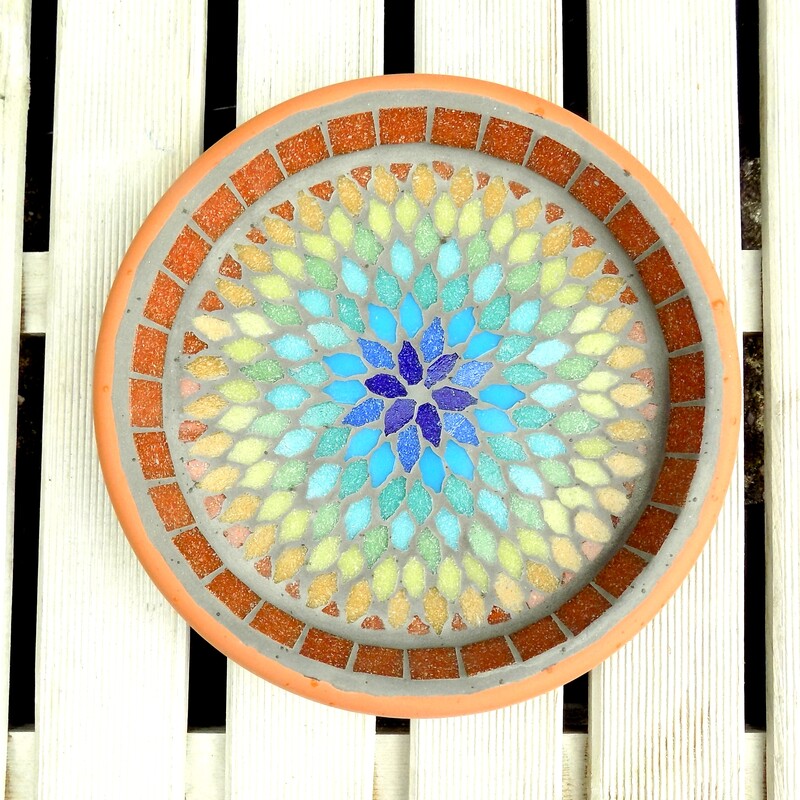 A mosaic garden  birdbath with a  mandala style design in the colours of the rainbow from terracotta on the outside to indigo blue in the centre