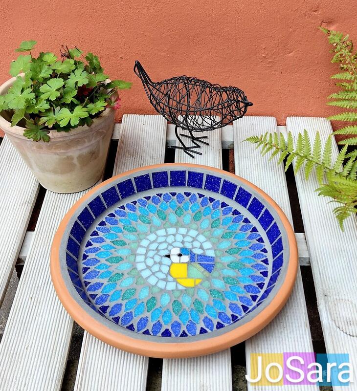 A mosaic garden birdbath with a bluetit bird in the centre of a splash effect made with blues, turquoise and greens.