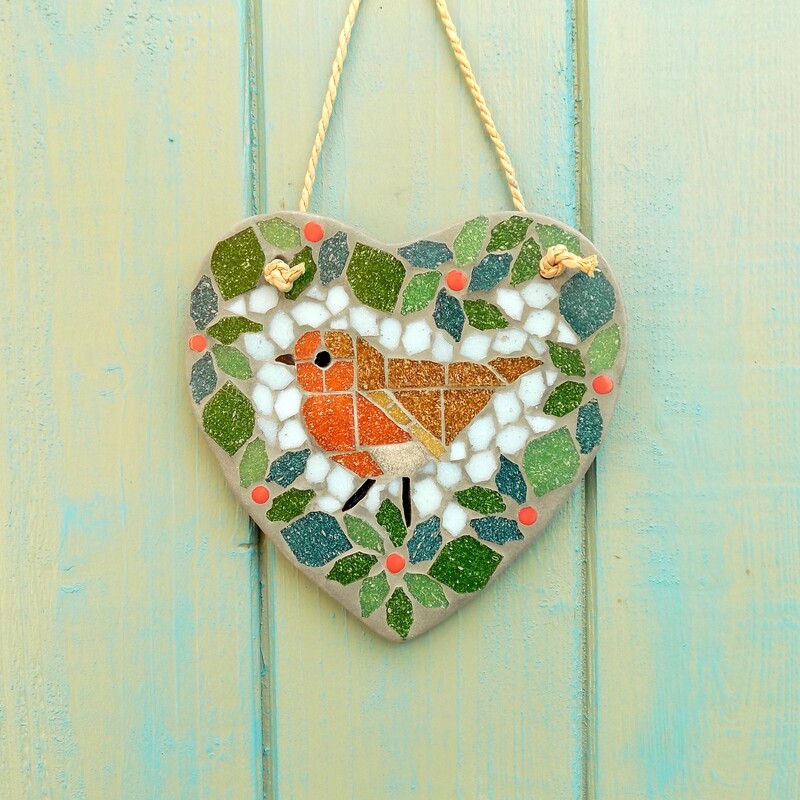 A mosaic garden hanging heart with a design of a robin bird standing in the centre of a foliage wreath with red berries