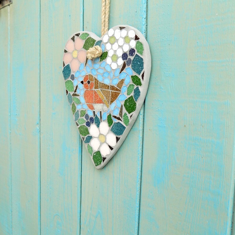 A handmade mosaic garden hanging heart with a design of a robin standing in a border of hedgerow leaves and flowers