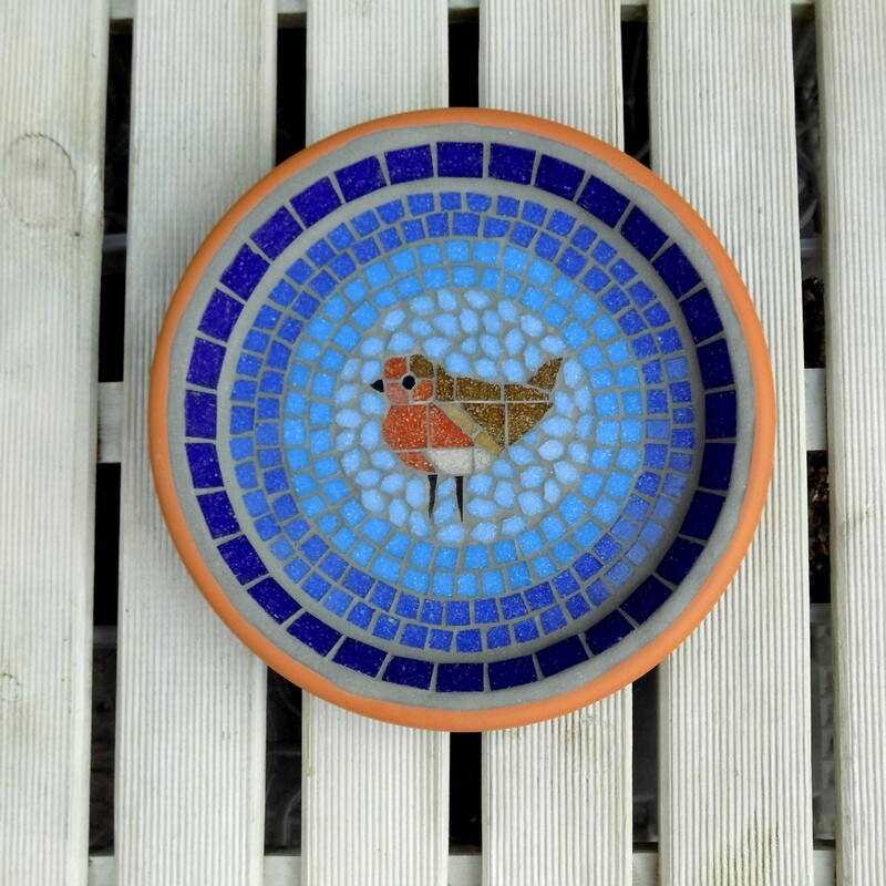 A mosaic garden bird bath with a robin bird in the centre of graduating shades of blue rings from dark on the border to a light sky blue around the bird