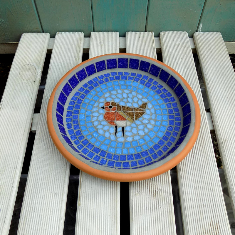 A mosaic bird bath with a robin bird in the centre of graduating shades of blue rings from dark on the border to a light sky blue around the bird