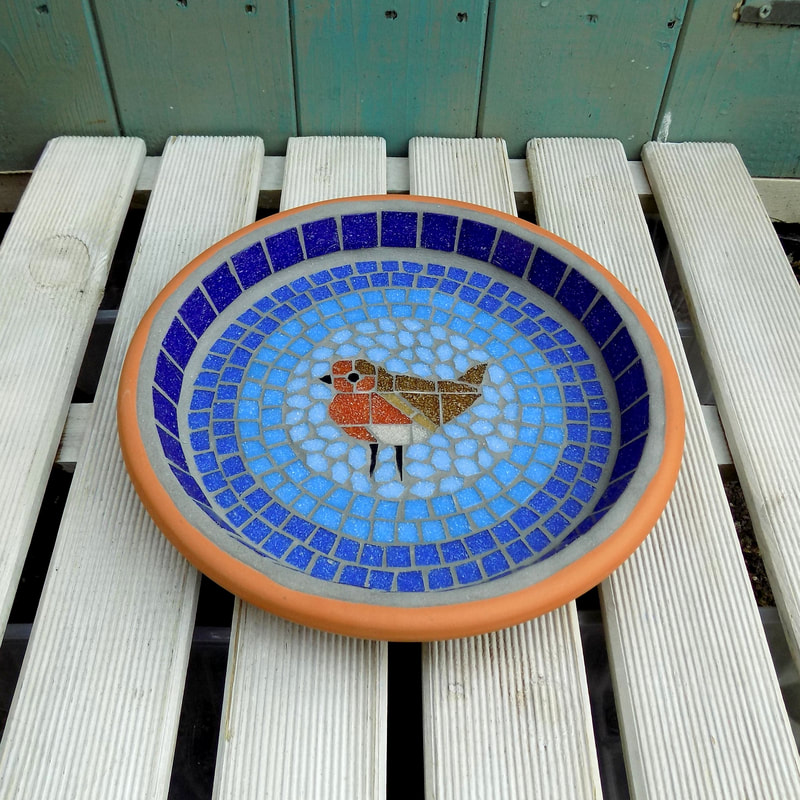 A mosaic garden birdbath with a robin bird in the centre of graduating shades of blue rings from dark on the border to a light sky blue around the bird