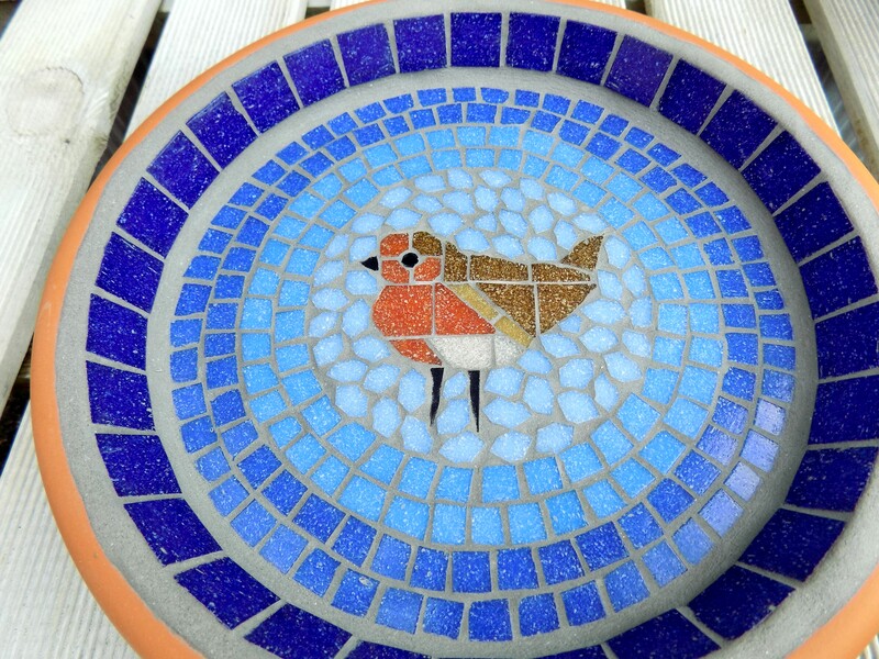 A mosaic birdbath with a robin bird in the centre of graduating shades of blue rings from dark on the border to a light sky blue around the bird