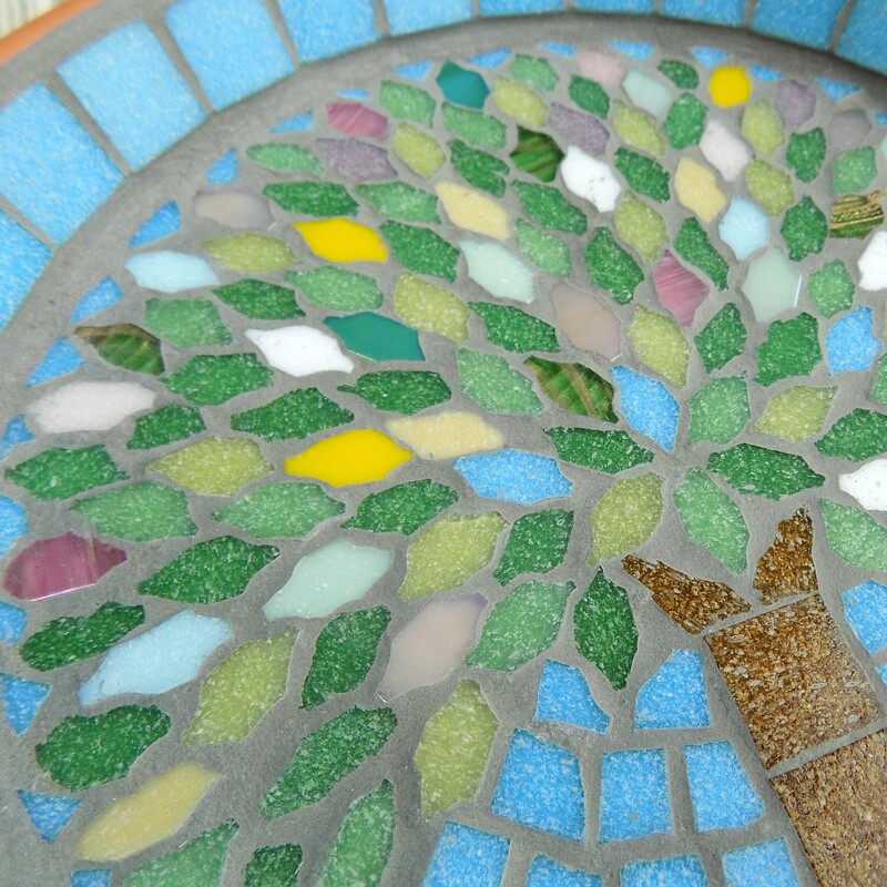 A mosaic bird bath with a design with a spring tree made using spring coloured tiles as the leaves and on a turquoise blue sky background