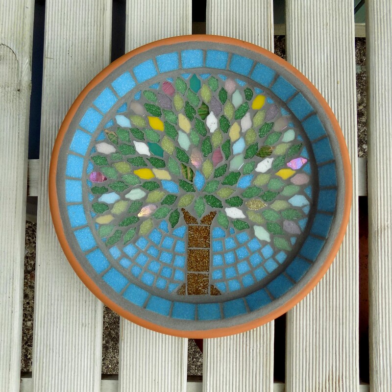A mosaic birdbath with a design with a spring tree made using spring blossom and fresh green tiles as the leaves and on a turquoise blue sky background