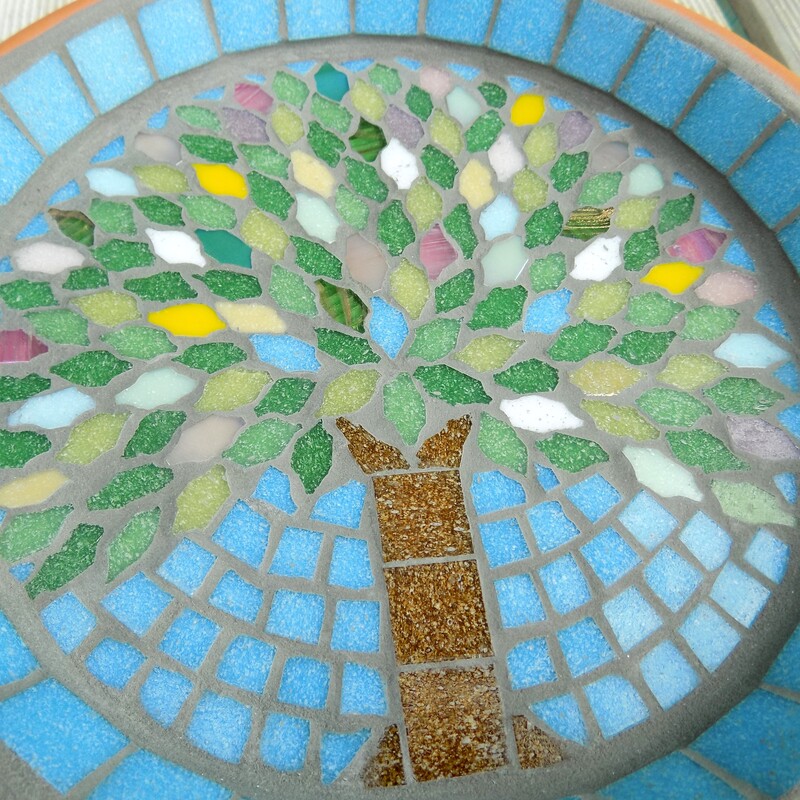 A mosaic garden bird bath with a design with a spring tree made using spring coloured tiles as the leaves and on a turquoise blue sky background