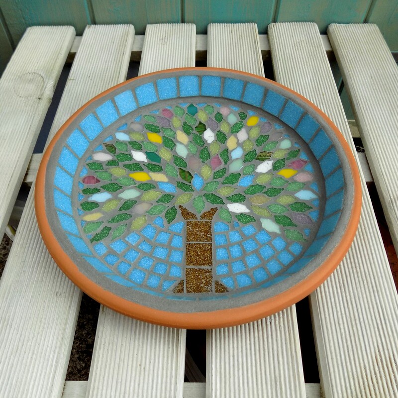 A mosaic garden birdbath with a design with a spring tree made using spring coloured tiles as the leaves and on a turquoise blue sky background