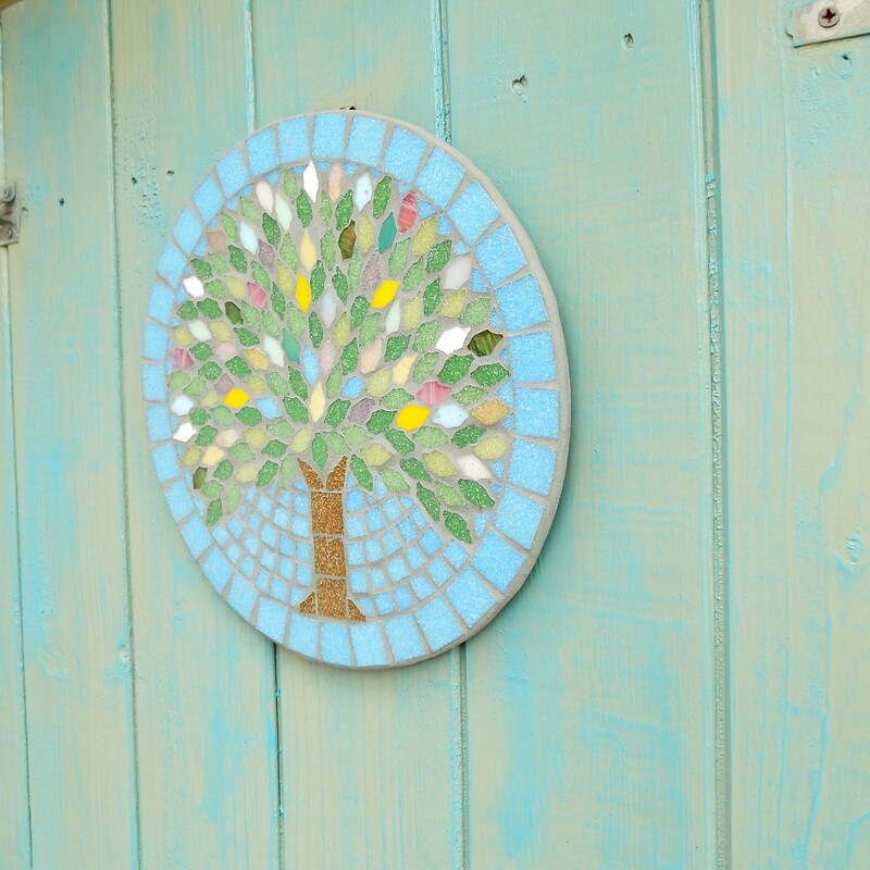 A mosaic garden hanging plaque with a design of a spring tree with leaves made in the colours of spring blossom and flowers on a turquoise blue background